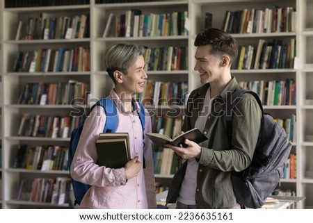 Two happy classmates student guys meeting in college library, standing together, reading open book, talking, laughing, discussing class project, homework tasks, education