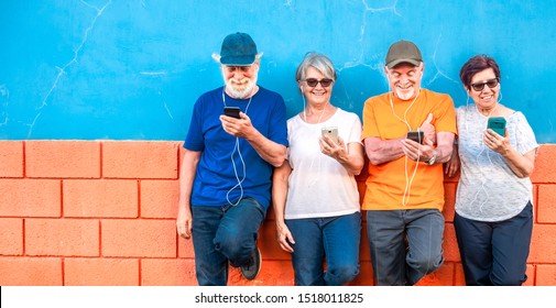 Two Happy Couples Of Senior People Standing Against An Orange And Blue Wall Smiling And Looking At The Mobile Phone Using Earphones. Casual Clothing And Relaxed Faces