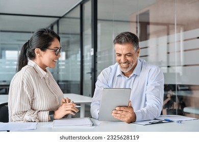 Two happy diverse executives team discussing work using digital tablet in office. Smiling Latin male manager talking to female Asian colleague working together on business finance project at meeting. - Powered by Shutterstock