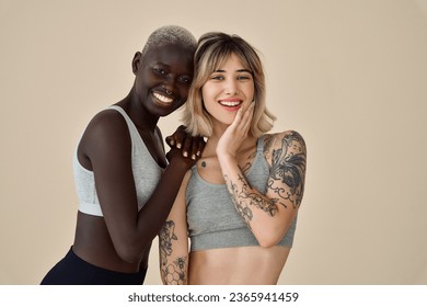 Two happy diverse fit women, Black and European young sporty gen z girls friends multiethnic models wearing sportswear tops standing smiling looking at camera on beige background, portrait.