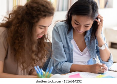Two happy diverse girls college friends study together in campus, teen asian and caucasian female students team talking laughing helping in preparing university creative research project make notes