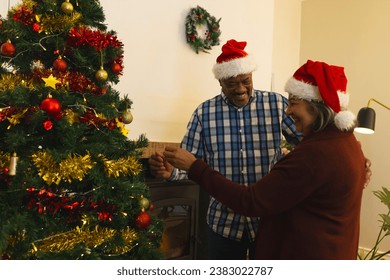 Two happy diverse male and female senior friends in santa hats, decorating christmas tree at home. Retirement, christmas, celebration, friendship, togetherness and senior lifestyle, unaltered. - Powered by Shutterstock