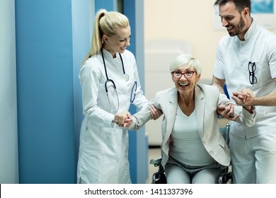 Two Happy Doctors Assisting Mature Patient To Get Up From The Wheelchair And Walk Again After Recovery. 