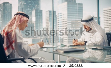 Two Happy Emirati Businessmen in White Traditional Kandura Sitting in Office and Signing Contract. Business Partners Sign Lucrative Investment. Saudi, Emirati, Arab Businessman Concept.