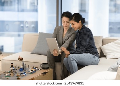 Two happy engaged diverse businesswomen using one tablet for Internet communication, sitting on couch in office lounge zone, discussing teamwork, online project, watching job content - Powered by Shutterstock