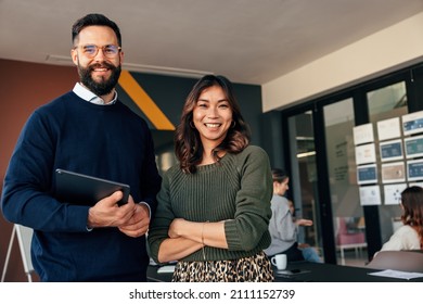 Two happy entrepreneurs smiling at the camera cheerfully. Young businesspeople standing in a boardroom with their colleagues in the background. Diverse entrepreneurs working together as a team. - Shutterstock ID 2111152739