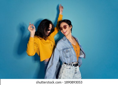 Two happy excited pretty stylish cool diverse gen z girls friends wearing glasses, trendy sunglasses and outfits having fun at hen night club party event, dancing together isolated on blue background.