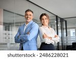 Two happy executive managers, mature business man and woman leaders, successful partners professional team in their 50s standing arms crossed in office at work looking at camera. Corporate portrait.