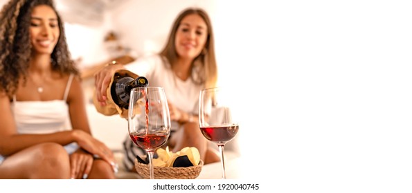 Two happy female friends celebrating at home pouring red wine in the glasses in selective focus effect and copy space. Young Caucasian woman toasting with her best Hispanic friend drinking alcohol - Powered by Shutterstock