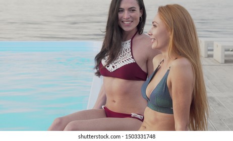 Two Happy Female Friends Chatting While Resting At The Poolside Together