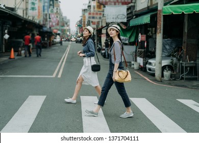 Two Happy Female Friends Travelers With Bags Crossing Street Together Outdoor Sunny Day In China Town. Japanese Lady Travel In Chinese City Walking On Zebra Cross In Urban. Girl Wear Hats Look At Sky.