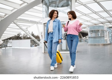 Two Happy Female Friends Travelling Together, Walking With Luggage At Airport, Smiling Young Women Carrying Suitcase And Chatting While Going To Flight Gate Through Terminal Hall, Copy Space - Powered by Shutterstock