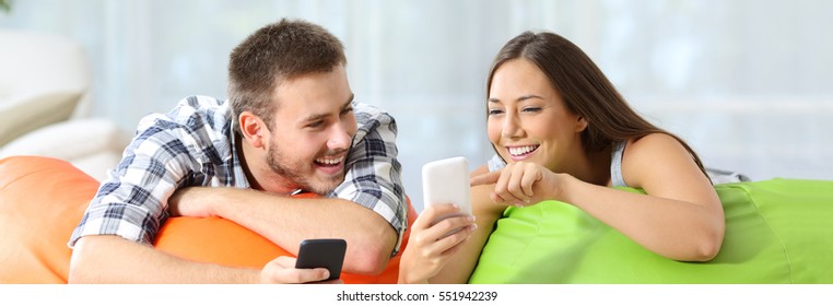 Two happy friends sharing media content on line with their smart phones lying on colorful puff in a house interior