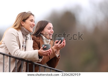 Two happy friends talking and drinking in a balcony in winter