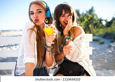 Two Happy Funny Hipster Girls Having Fun On Beach Summer Party, Laughing And Sensing Kiss, Drinking Tasty Cocktail, Bright Summer Outfits And Accessories, Listening Music At Headphones.