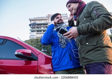 Two Happy Gay Guys Listening Music From Mobile By Sharing Headphones Next To Red Car In The City. LGBTQ People Life. Copy Space.	