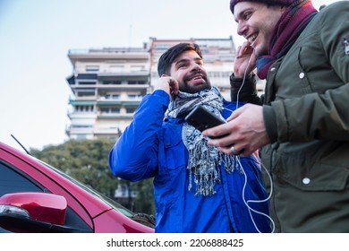 Two Happy Gay Guys Smiling While Listening Music From Mobile And Sharing Headphones Next To Red Car In The City. LGBTQ People Life. Copy Space.	