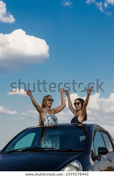 Two happy girlfriends are enjoying a car\
ride. Two beautiful blonde and brunette girls look out of the hatch\
of the car and enjoy life. Vertical\
photo