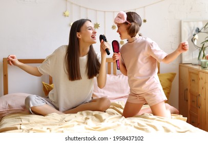 Two Happy Girls Pretending To Be Famous Singers, Overjoyed Younger And Older Sister With Hairbrush As Mic Singing While Sitting On The Bed. Young Mom And Cute Little Daughter Having Fun At Home