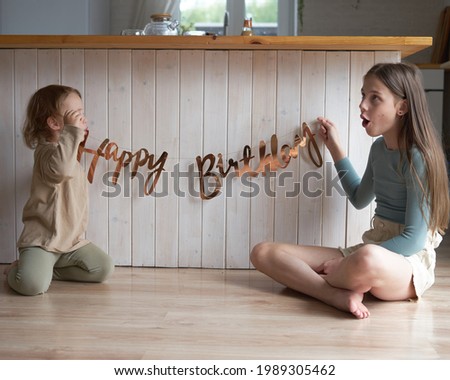 two happy girls sit at home in the afternoon on the floor holding the inscription happy birthday and fooling around                               