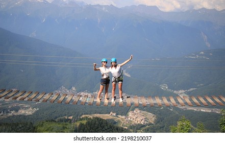 Two Happy Girls Stand On A Rope Bridge In The Mountains. Extreme Vacation.