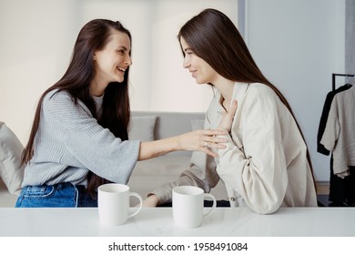 Two happy girls talking to each other, sitting together on kitchen, drink beverage and spending weekend at home. Smiling girl telling unbelievable story to her best friend while she laugh