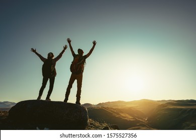 Two happy hikers stands with raised arms against sunset and enjoys mountains view