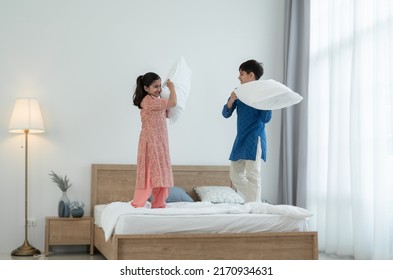 Two happy Indian brother and sister in traditional clothing standing on bed, playing pillow fight, having fun together at home. Playful kids, Siblings relationship concept