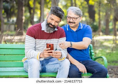 Two Happy Indian Senior Men Watching Video Or Social Media On Smart Android Phone While Sitting At Park Outdoor, Mature Old People Using Technology, Friends Having Fun.