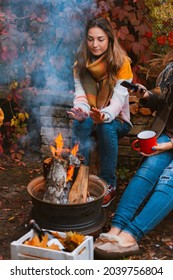 Two Happy Joyful Best Friends Sitting Outside In Autumn Garden By Campfire With Bottle Of Red Wine