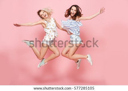 Two happy joyful young women jumping and laughing together over pink background