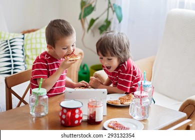 Two Happy Kids, Two Brothers, Having Healthy Breakfast Sitting At Wooden Table In Sunny Kitchen, Eating Waffles  And Watching Cartoon On Tablet