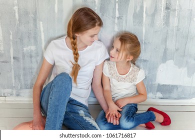 Two happy kids sitting against grey textured wall background and embracing. Adorable pretty little girl hugging tight cute teenage girl, showing her love and care. Sisters having fun at home
