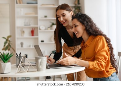 Two Happy Ladies Using Laptop Computer Working And Learning Online Together Or Making Video Call Sitting At Desk In Modern Office. E-Learning. Females Entrepreneurship Career Concept. Side View