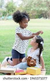 Two Happy Little Diverse Kids, African And Caucasian Girls Helping Stick Hairpin On Her Friend's Hair With Love While They Picnic Playing Together In Green Park. Diversity Ethnic Friendship Concept