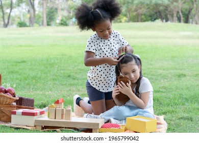 Two Happy Little Diverse Kids, African And Caucasian Girls Helping Stick Hairpin On Her Friend's Hair With Love While They Picnic Playing Together In Green Park. Diversity Ethnic Friendship Concept