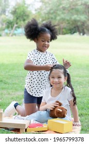 Two Happy Little Diverse Kids, African And Caucasian Girls Helping Stick Hairpin On Her Friend's Hair With Love While They Picnic Playing Together In Green Park. Diversity Ethnic Friendship Concept