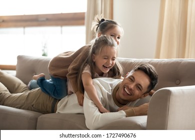 Two Happy Little Girls Playing Lying On Couch With Father, Smiling Young Dad Having Fun With Preschool Daughters, Laughing, Relaxing On Cozy Sofa At Home, Funny Family Activity, Enjoying Weekend