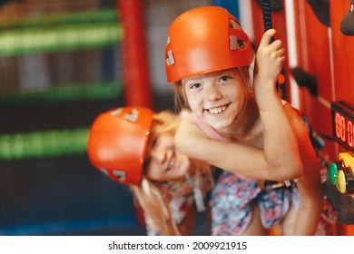 Two Happy Little Girls In Red Helmets Climbing The Wall In Bouldering Center. School Girls Smiling At The Camera And Having Fun In Indoor Playground For Children. Indoor Climbing Class For Kids