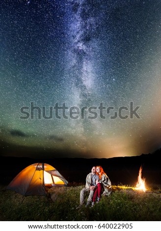 Two happy lovers covered with a plaid sitting together near campfire and camp at night under stars and looking to the starry sky and Milky way