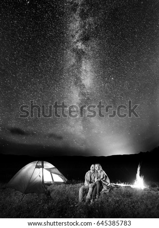 Two happy lovers covered with a plaid sitting together near campfire and camp at night under stars and looking to the starry sky and Milky way. Black and white