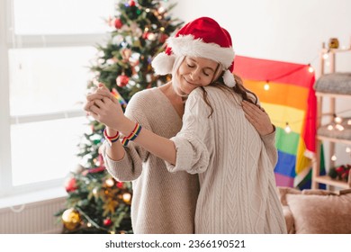 Two happy loving lesbian women celebrate Christmas or New year at home. Middle-aged women dance in Christmas hats at the rainbow LGBT flag - Powered by Shutterstock