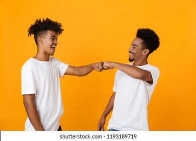 Two Happy Male African Friends In T-shirts Standing Isolated Over Yellow Background, Giving Fist Bump