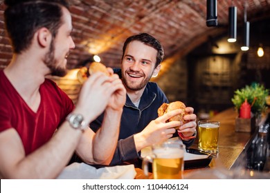 Two Happy Male Friends Having Dinner At Popular Burger Bar. Young People Sitting, Talking And Eating Burgers, Drinking Beer.