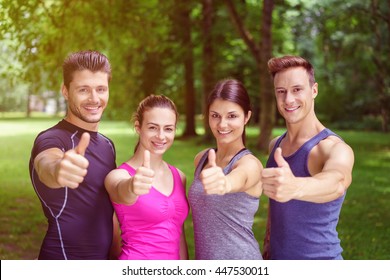 Two happy motivated fit young couples giving a thumbs up of approval and success and they pose in their sportswear in a park smiling at the camera - Powered by Shutterstock