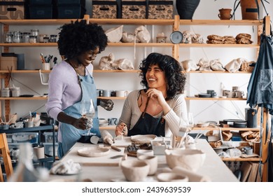 Two happy multicultural craftswomen making earthenware and drinking wine at pottery workshop in studio. Cheerful interracial women are laughing and having fun with their hobby in pottery workshop. - Powered by Shutterstock