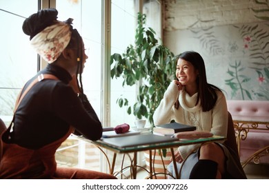 Two Happy Multicultural Female College Girls Sit In The Cafeteria, Chatting And Gossiping. Female Students And Girls Talk.