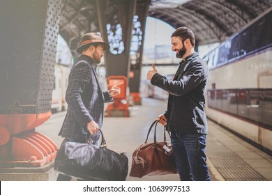 Two happy multi-ethnic bearded businessmans together wearing casual clothes and holding travel bags in hands. Friends casual talking while waiting the train on the railway platform.