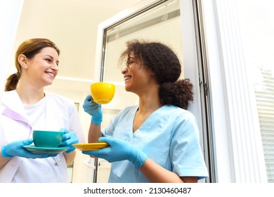 Two happy multi-ethnic female medical workers colleagues drinking coffee or tea and talking while standing near window in modern clinic or hospital, taking a break from work - Powered by Shutterstock