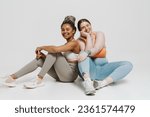 Two happy multiethnic women with different body types in sportswear posing together while sitting isolated over white wall
